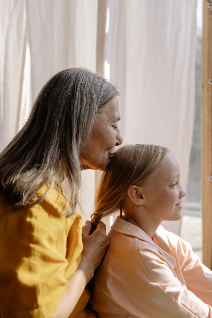 Elderly woman lovingly braiding young girl's hair by sunlit window, emphasizing family bond.